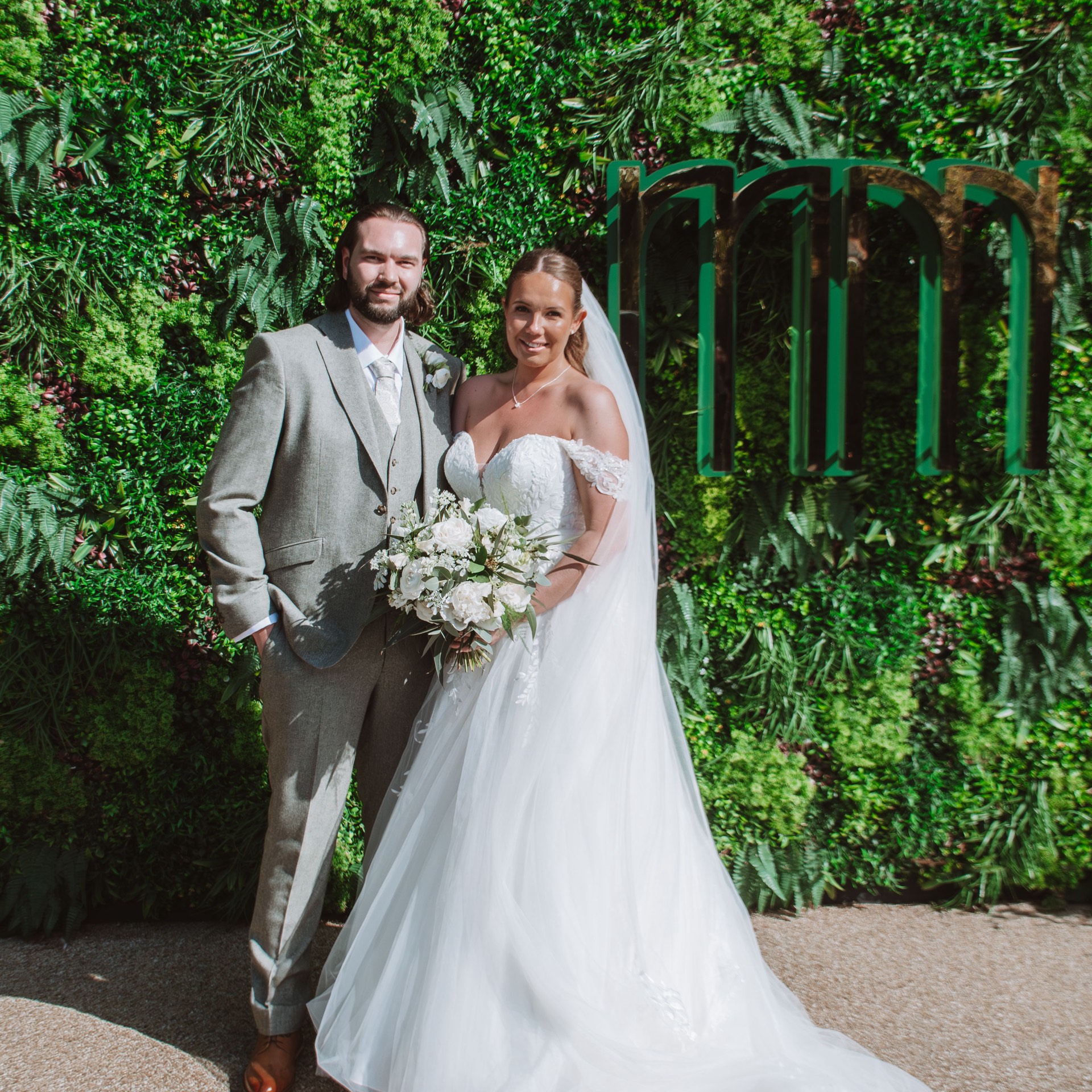 A bride and groom posing together outdoors in front of a green wall