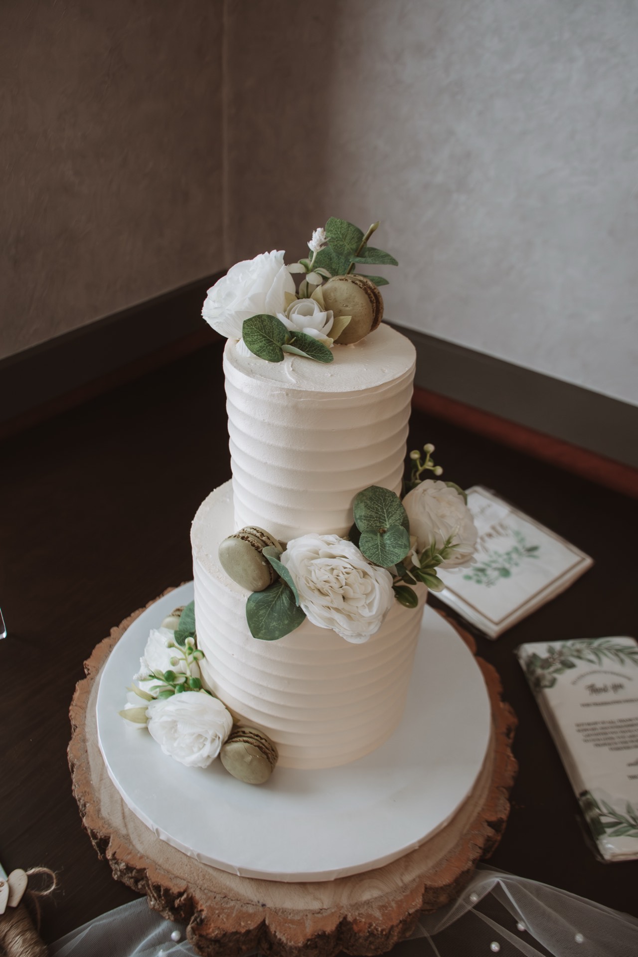 A two-tiered white wedding cake with flowers, leaves and macarons
